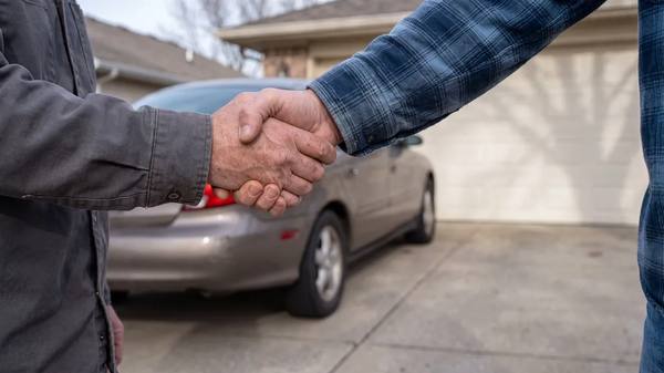 Two people shaking hands beside an older sedan parked on a driveway