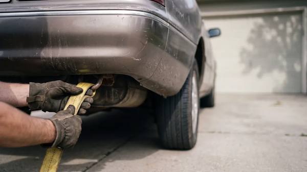 Work-gloved hands hooking a yellow recovery strap to the rear tow point of an older car on a driveway