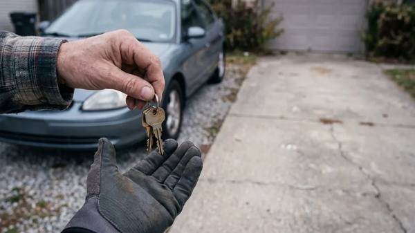 A set of car keys passed from a bare hand into a work-gloved hand, beside an older sedan parked on a residential driveway